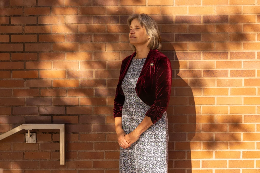 A woman with a red velvet blazer and patterned dress stands in front of a brick wall for a portrait